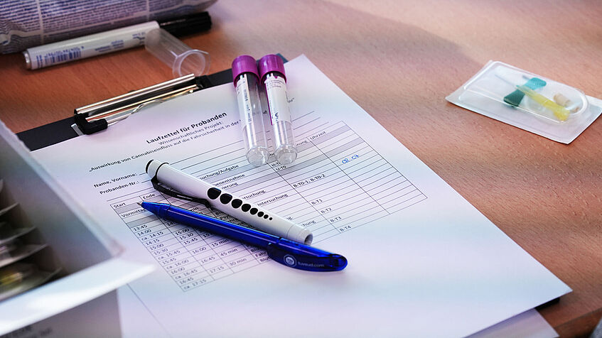 Ein Blattpapier mit einer Excel-Tabelle und Stiften liegt auf einem Tisch. Daneben liegen noch verpackte Utensilien für die Blutentnahme. 