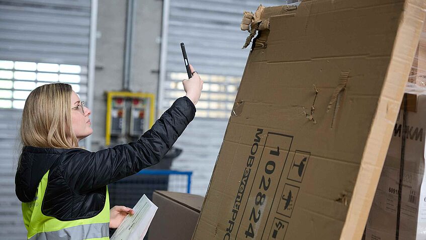 Tessa Erzgräber fotografiert eine beschädigte Verpackung mit dem Smartphone im Logistiklager.