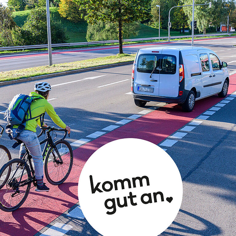 Zwei Radfahrende fahren auf einem rot markierten Radweg, ein Lieferwagen fährt voraus und blinkt, weil er die Spur wechselt und dabei den Radweg überfährt, städtische Umgebung mit Bäumen und Straßenmarkierungen.