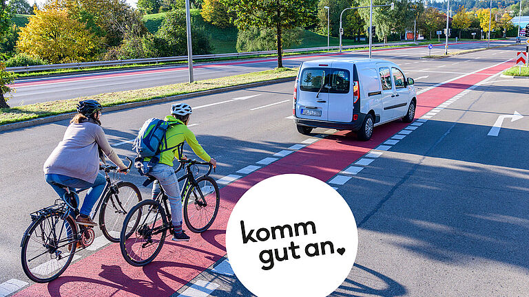 Zwei Radfahrende fahren auf einem rot markierten Radweg, ein Lieferwagen fährt voraus und blinkt, weil er die Spur wechselt und dabei den Radweg überfährt, städtische Umgebung mit Bäumen und Straßenmarkierungen.