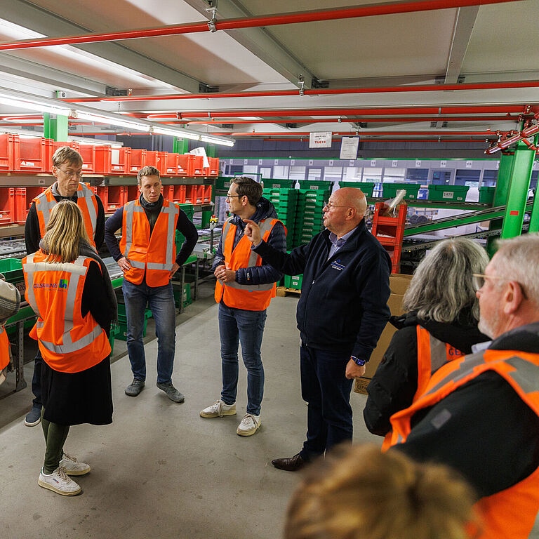 Eine Gruppe von Menschen steht im Lager eines Logistikbetriebes. Alle tragen leuchtend rote Warnwesten. Außer einem Mann in blauer Jacke, der Betriebsleiter. Er erklärt die Arbeitsabläufe.. Im Hintergrudn sind 