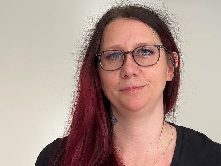 Ergotherapeutin Iris Stenzel im Porträt: Sie schaut freundlich in die Kamer. Die Brillenträgerin hat lange rote Haare. Am rechten Arm sieht man eine Tätowierung. 