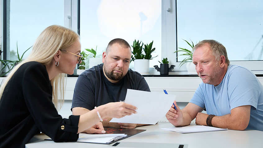 Drei Personen sitzen an einem Tisch, betrachten gemeinsam Dokumente und besprechen etwas konzentriert in einem hellen Büro mit Pflanzen am Fenster.