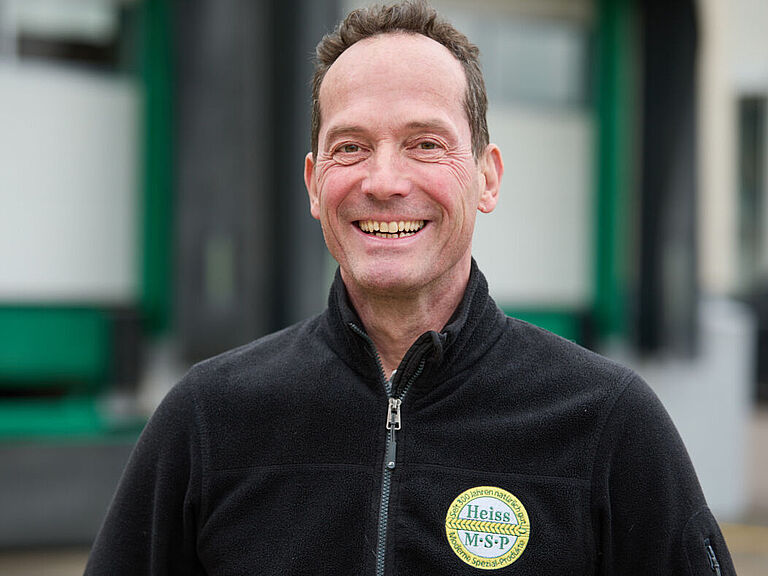 Ein Mann in schwarzer Arbeitsjacke schaut lachend in die Kamera.