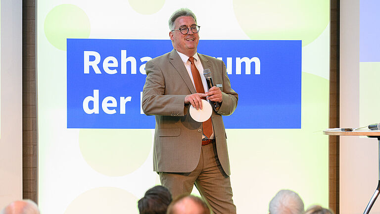 Martin Kögler, steht auf der Bühne des Reha-Forums und spricht zum Publikum, das von hinten zu sehen ist. Er trägt einen hellbraunen Anzug. Hinter ihm leuchtet der Schriftzug Reha-Forum. 