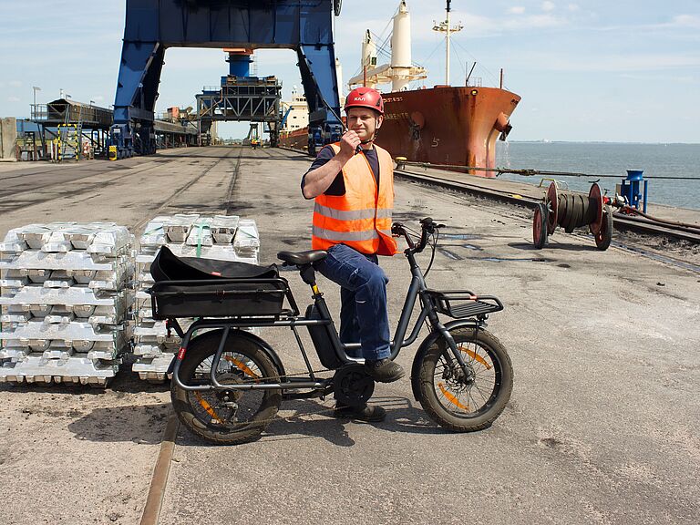 Ein Hafenarbeiter von Brunsbüttel Ports mit orangefarbiger Warnweste und rotem Helm sitzt auf einem schwarzen Lasten-E-Bike und hält ein Funkgerät ans Ohr. Neben ihm liegen gestapelte Metallblöcke, im Hintergrund sind ein großer Kran und ein Frachtschiff am Kai zu sehen.