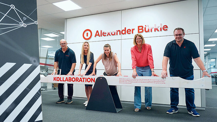 Fünf Personen stehen hinter eine Wippe in einem Büro