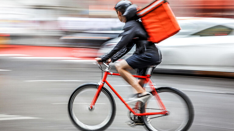 Ein Radfahrer fährt mit einem roten Mountain-Bike und einem roten Lieferrucksack über eine Straße. Im Hintergrund sieht man verwischte Autos. Der Radfahrer trägt Helm, kurze Hosen und eine schwarze Jacke. 