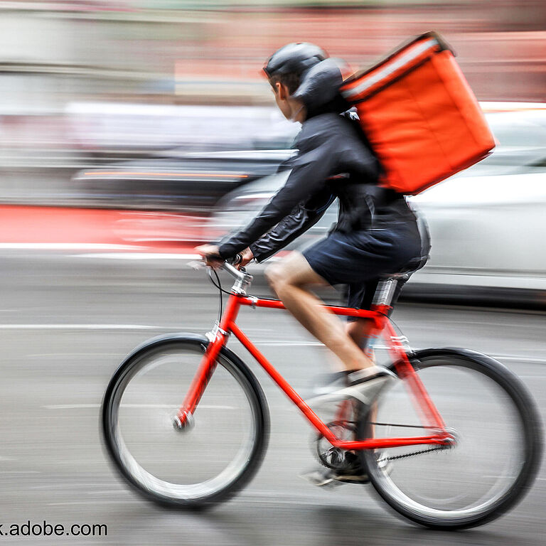 Ein Radfahrer fährt mit einem roten Mountain-Bike und einem roten Lieferrucksack über eine Straße. Im Hintergrund sieht man verwischte Autos. Der Radfahrer trägt Helm, kurze Hosen und eine schwarze Jacke. 