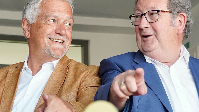 Zwei Männer sitzen auf einem Schreibtisch, Schulter an Schulter, schauen sich lächelnd an und zeigen auf das Logo der Deutschen Gesetzlichen Unfallversicherung, das vor ihnen auf dem Tisch steht.