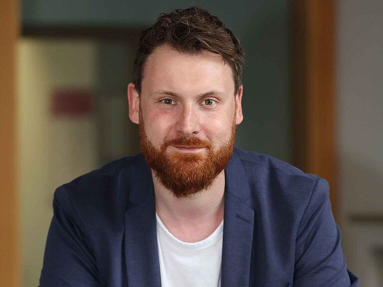 Alexander Tjaberings, ist Referent im Dezernat Einwirkungen und Berufskrankheiten der BGHW, lehnt an einem Geländer und lächelt in die Kamera. 