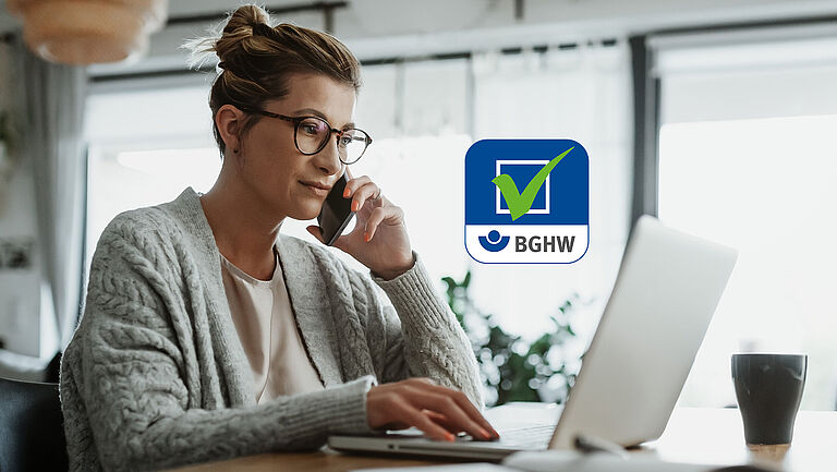 Eine Frau sitzt in ihrem heimischen Wohnzimmer an einem Laptop und telefoniert. Im Bild mittig ist das Logo der Gefährdungsbeurteilung Online der BGHW zu sehen.