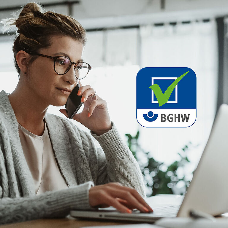 Eine Frau sitzt in ihrem heimischen Wohnzimmer an einem Laptop und telefoniert. Im Bild mittig ist das Logo der Gefährdungsbeurteilung Online der BGHW zu sehen.