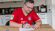 Steven Wedhorn sitzt in seinem roten Trikot am Tisch und schreibt eine Einkaufsliste. Hinter ihm sieht man Küchenschränke. 