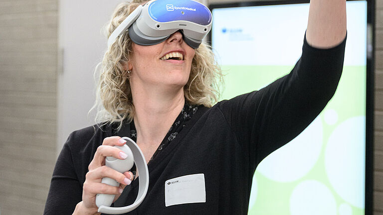 Eine Frau Trägt eine VR-Brille, eine Virtual-Reality-Brille und schaut auf einen Gegenstand, den sie in der linken Hand hält. 