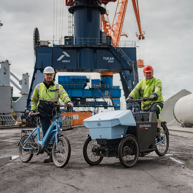 Zwei Hafenmitarbeiter in Warnschutzkleidung nutzen Fahrräder auf dem Hafengelände von Brunsbüttel Ports. Links steht ein Mann mit Helm neben einem kompakten blauen Fahrrad, rechts fährt ein Kollege mit rotem Helm ein dreirädriges Lastenrad mit großer Transportbox. Im Hintergrund sind Kräne, Container und ein großes Bauteil sichtbar.
