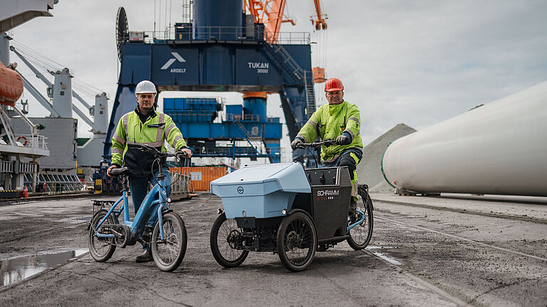 Zwei Hafenmitarbeiter in Warnschutzkleidung nutzen Fahrräder auf dem Hafengelände von Brunsbüttel Ports. Links steht ein Mann mit Helm neben einem kompakten blauen Fahrrad, rechts fährt ein Kollege mit rotem Helm ein dreirädriges Lastenrad mit großer Transportbox. Im Hintergrund sind Kräne, Container und ein großes Bauteil sichtbar.