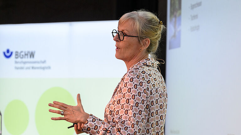 Dieses Bild zeigt die Leitende Oberärztin der BG Klinik Murnau, Sabine Drisch, in ihrem Vortrag im BGHW-Reha-Forum. Sie trägt eine bunte Bluse, steht seitlich zur Kamera und gestikuliert. 