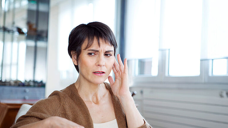 Eine Frau sitzt in einem Büro und tastet mit der linken Hand am linken Ohr, ihr Gesichtsausdruck ist unentspannt.