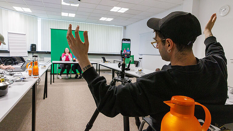 Dreharbeiten: Im Vordergrund hebt ein Mann die Hände, im Hintergrund drehen junge Menschen vor einem Greenscreen