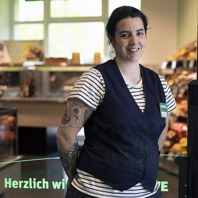 Eine Frau im gestreiften T-Shirt und blauer Weste steht am Eingang eines Lebensmittelmarktes und lacht freundlich in die Kamera. Im Hintergrund:bunte Gemüse- und Obststände. 