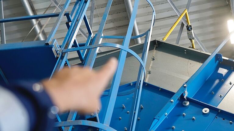 Eine Hand zeigt sich nach oben auf einen grauer Kasten an einem gelben Metallrahmen, der sich über dem Einfülltrichter der Müllpresse befindet. Von dort wird das Antennensignal gesendet. 