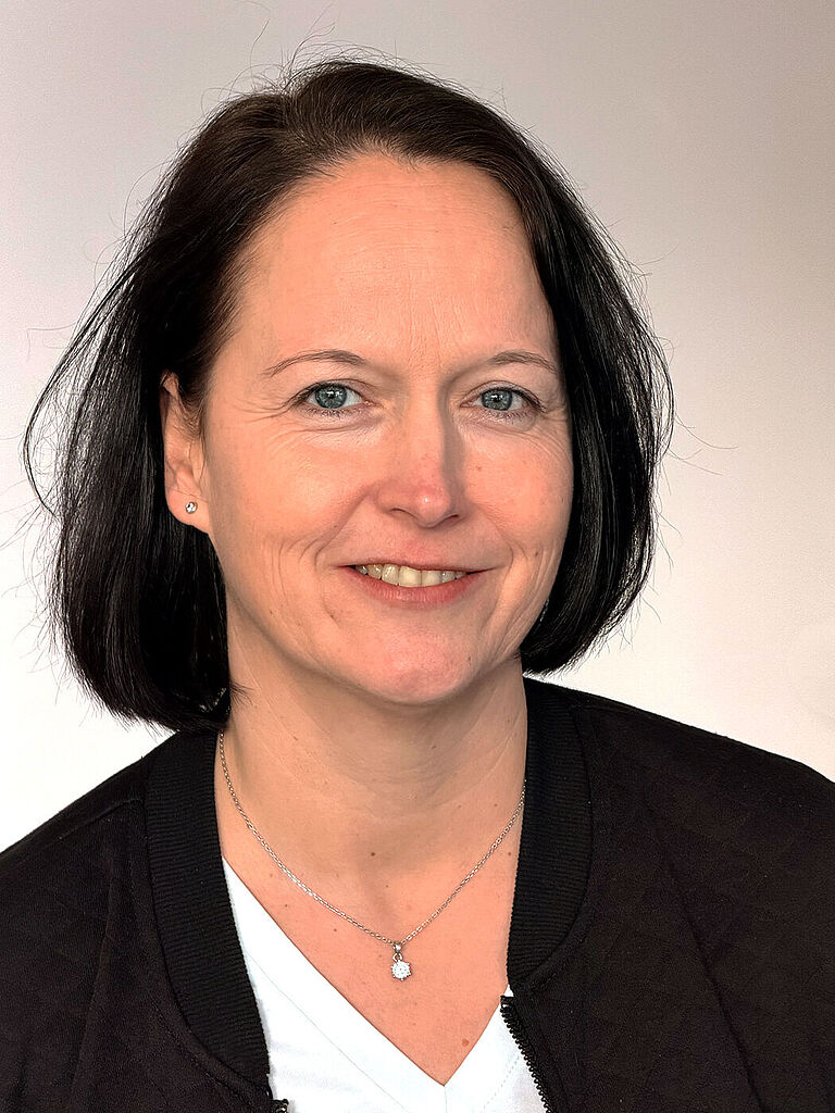 Ein Porträtfoto von Ina Papen, eine Frau mit Schulterlangen schwarzen Haaren, schaut freundlich in die Kamera. Sie traägt ein weißes T-Shirt und eine schwarze Jacke darüber. 