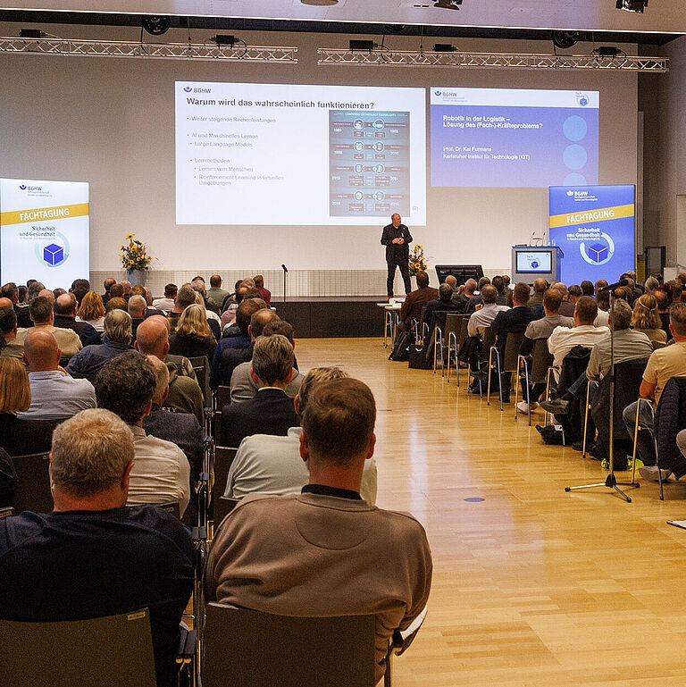 Eröffnung der BGHW-Fachtagung 2024 - Blick über vollbesetzte Stuhlreihen aufs Podium
