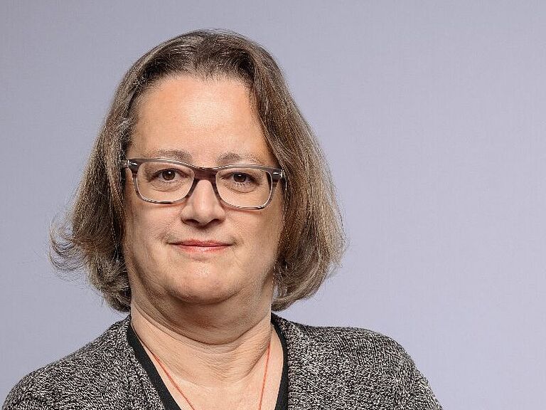 Portraitfoto einer Frau mit mittellangen braunen Haaren und Brille, die eine graue Strickjacke über einem schwarzen Oberteil trägt. Neutraler Hintergrund.
