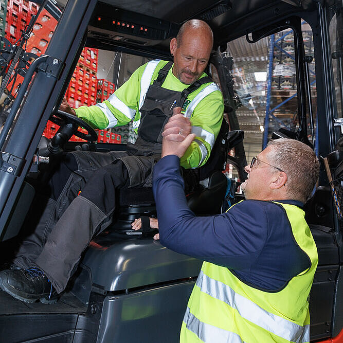 Ein Mann in Arbeitshose sitzt auf einem Gapelstapler, ein Mann im Jacket steht neben ihm. Beide schütteln sich die Hände als Zeichen der Verbundenheit. Beide tragen gelbe Warnwesten. 