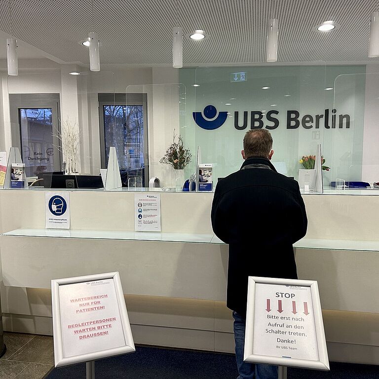 Im Foyer der Unfallbehandlungsstelle steht ein Mann im dunklen Mantel vor einem weißen Empfangsschalter. Im Hintergrund steht auf einer Glasscheibe UBS Berlin geschrieben. 
