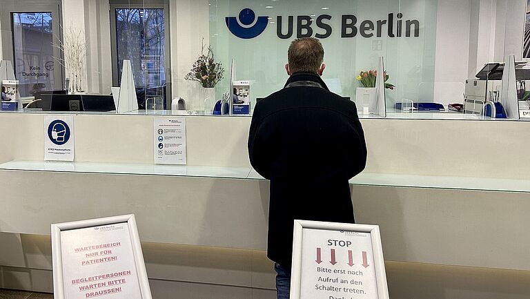 Im Foyer der Unfallbehandlungsstelle steht ein Mann im dunklen Mantel vor einem weißen Empfangsschalter. Im Hintergrund steht auf einer Glasscheibe UBS Berlin geschrieben. 