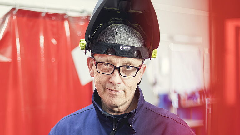 Udo Westerwick steht vor einer Rohrleitung, die er bearbeitet. Die Schutzhaube auf seinem Kopf hat er hochgeschoben. Er lacht in die Kamera.