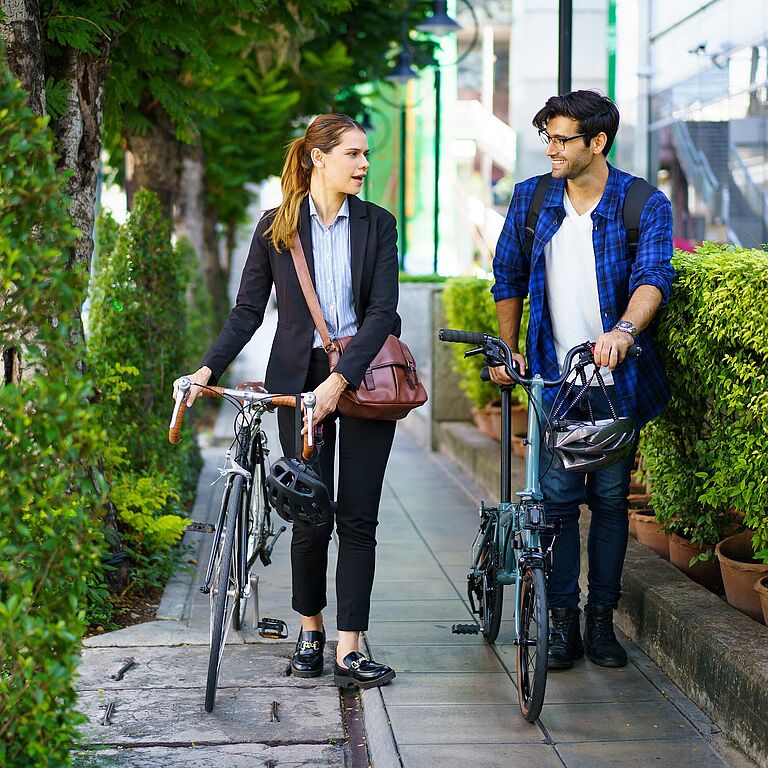 Eine Frau im Business-Outfit und ein Mann in Freizeitkleidung gehen nebeneinander auf einem Gehweg, während sie jeweils ihre Fahrräder schieben. Beide tragen Umhängetaschen, halten Fahrradhelme in der Hand und unterhalten sich lächelnd. Im Hintergrund stehen grüne Hecken und Bäume am Straßenrand.