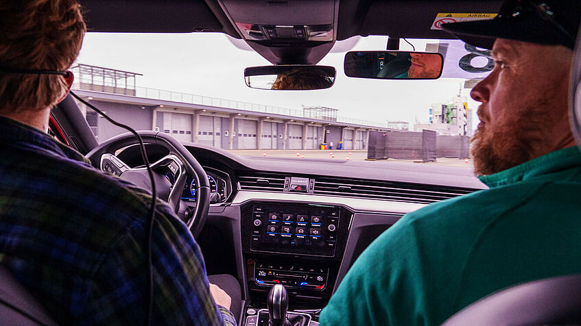 Blick von der Rückbank ins Pkw-Cockpit bei einer Testfahrt: Rechts sitzt ein Fahrlehrer, links daneben ein Person, die unter seiner Regie Fahrübungen durchführt. Vor ihnen liegt der Parcours, der auf dem Sachsenring abgesteckt wurde. 
