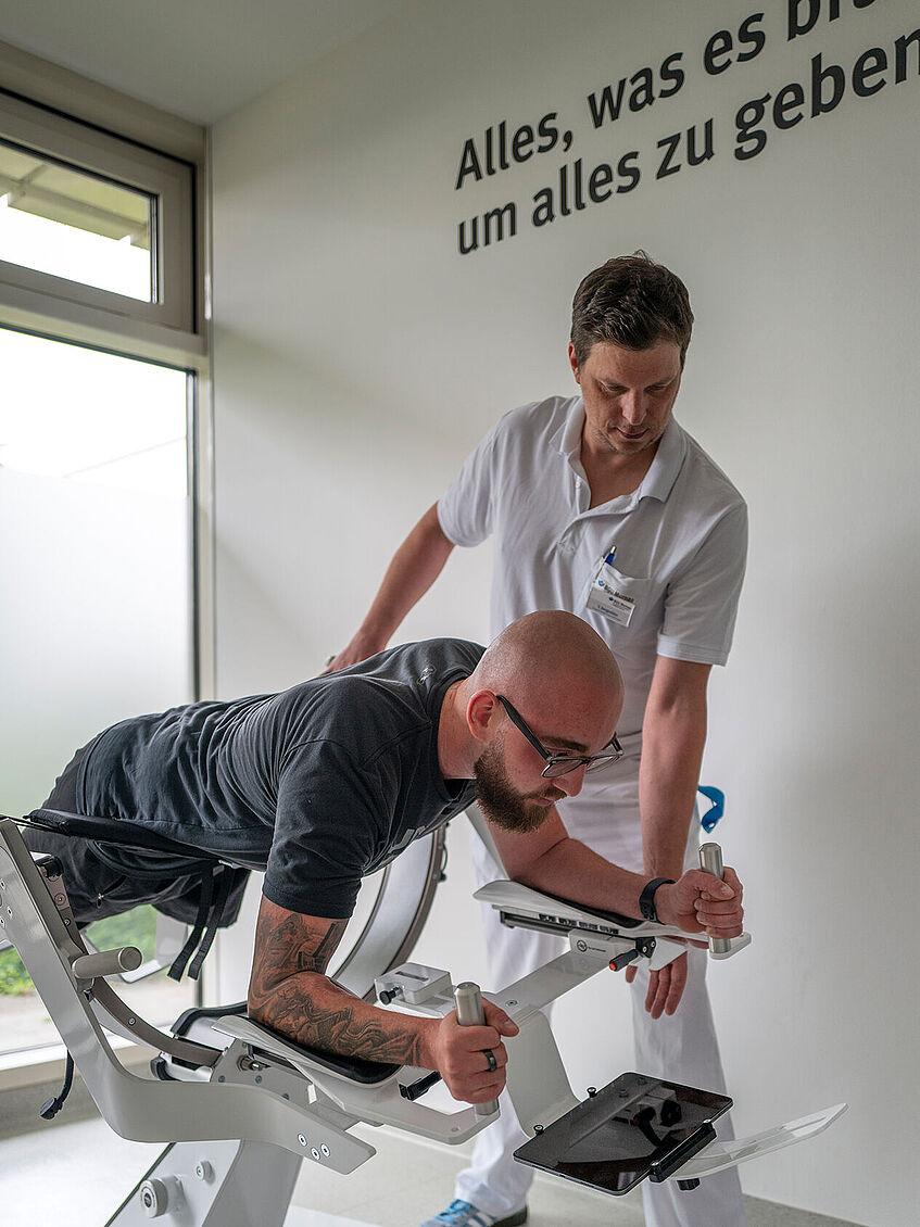 Moritz Sontheimer lieg in einem Spezialgerät für Stabilitätstraining, mit dem Gesicht zum Boden. Physiotherapeut Bergmüller steht hinter ihm und korrigiert die Haltung.