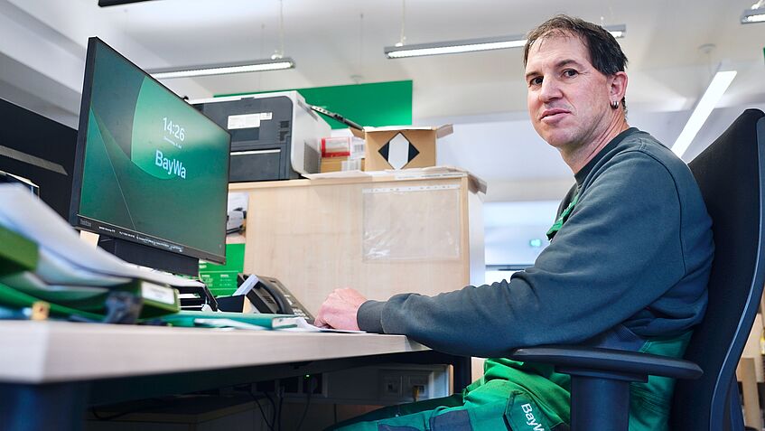 Ein Mann in Arbeitskleidung an einem Schreibtisch vor einem Computerbildschirm.