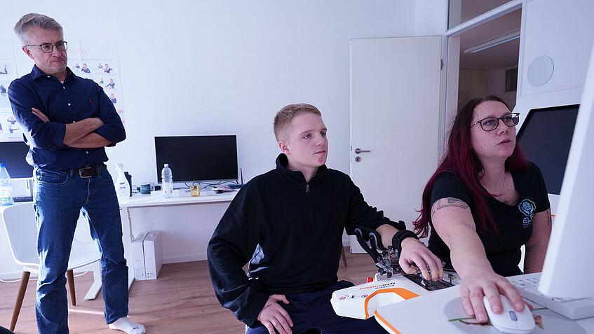 Handtherapie am Spezialgerät: Ergotherapeutin Iris Stenzel, Lars Eberhard (beide sitzend) und Reha-Berater Hartmut Wahlig schauen auf den Bildschirm des Therapie-Computers, an dem Lars seine Hände trainiert. 