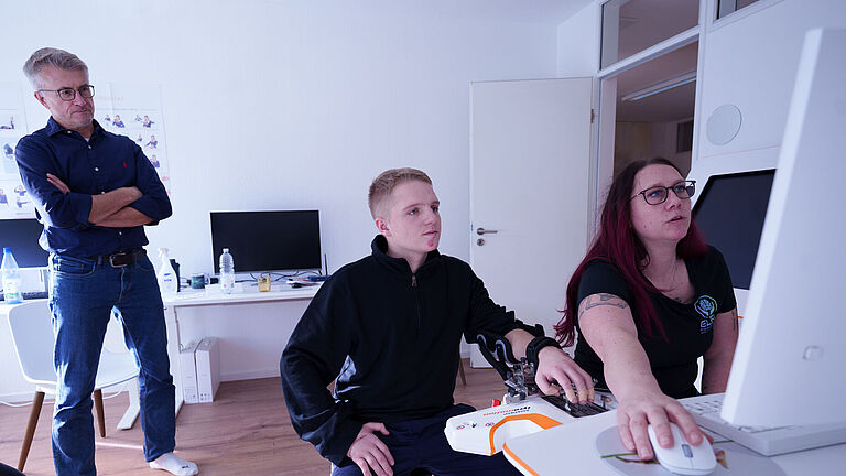 Lars und Iris Stenzel sitzen vor einem Computer. Im Hintergrund steht Reha-Berater Hartmut Wahlig. 