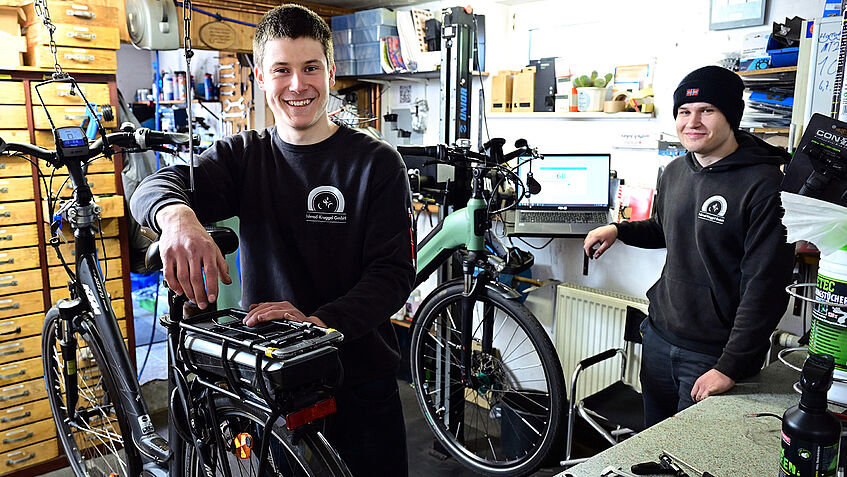 Zwei junge Männer in einer Fahrradwerkstatt, einer kontrolliert ein Akku an einem E-Bike.