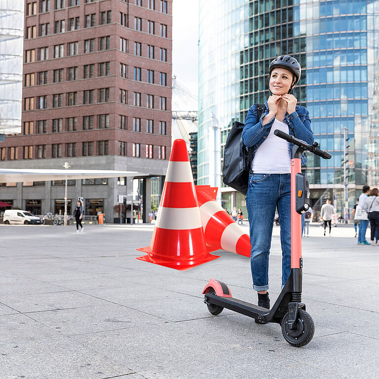 Eine Frau steht vor eine Hochhausfassade, neben ihr ein E-Scooter, während sie sich gerade einen Helm anzieht. Hinter ihr stehen große Pylone als Symbol für Fahrsicherheitstraining