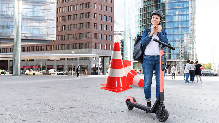 Eine Frau steht vor eine Hochhausfassade, neben ihr ein E-Scooter, während sie sich gerade einen Helm anzieht. Hinter ihr stehen große Pylone als Symbol für Fahrsicherheitstraining