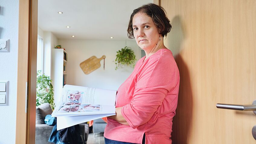 Eine Frau lehnt an einer Tür und hält eine Mappe mit Bildern in der Hand, Blick in die Kamera.