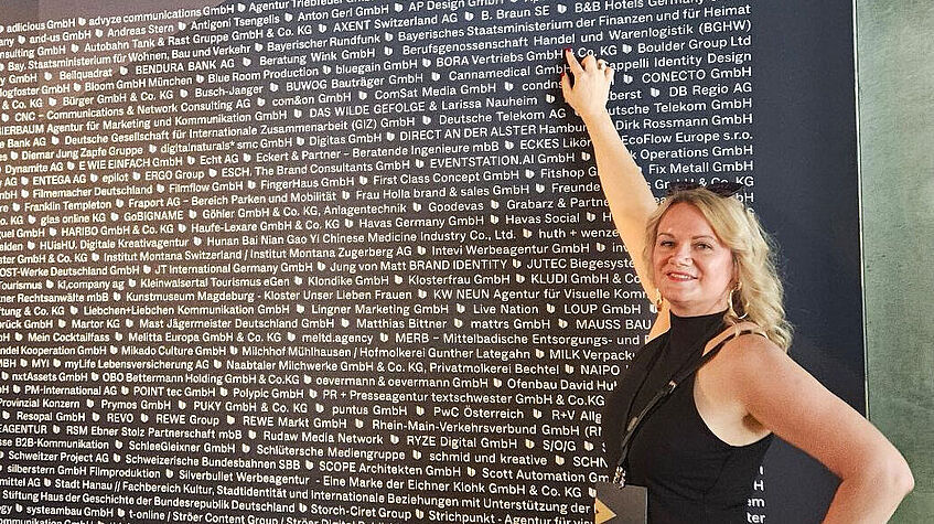 Eine Frau steht vor einer Wand mit vielen Firmennamen und zeigt auf den Namen BGHW