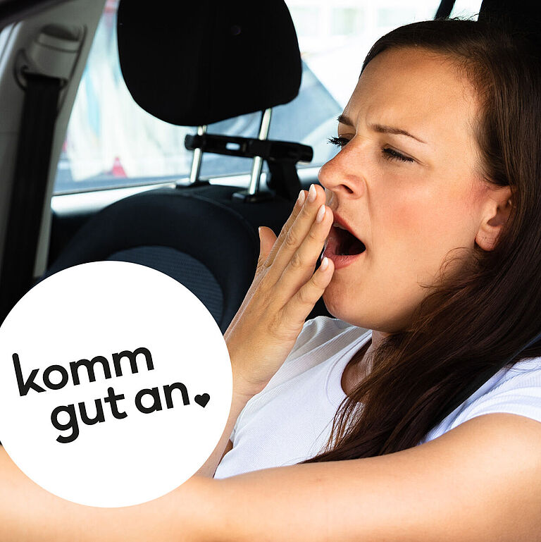 Junge Frau mit langen dunklen Haaren sitzt am Steuer eines Autos, gähnt und hält sich eine Hand vor den Mund. Auf dem Bild steht der Schriftzug ‚komm gut an‘.