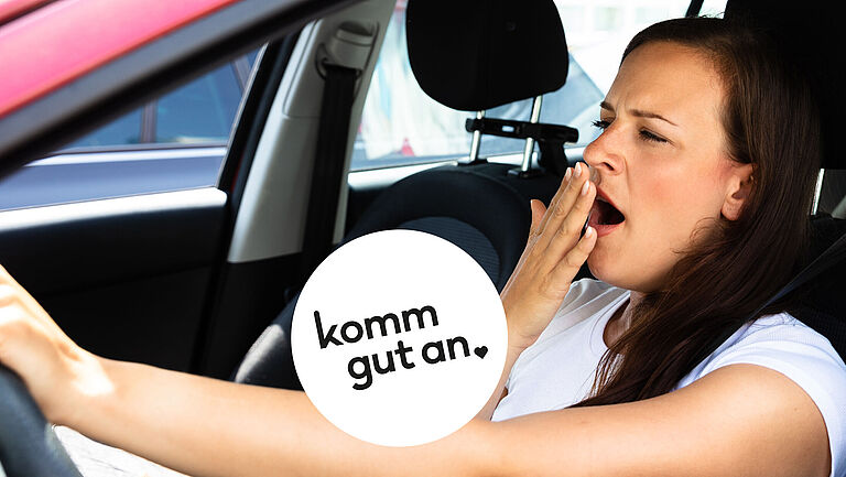 Junge Frau mit langen dunklen Haaren sitzt am Steuer eines Autos, gähnt und hält sich eine Hand vor den Mund. Auf dem Bild steht der Schriftzug ‚komm gut an‘.