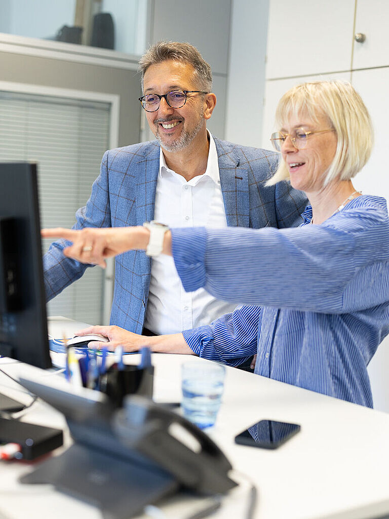 Karl-Josef Thielen mit blauem Sakko, an Schreibtisch bei blonder Mitarbeiterin. Beide schauen sich etwas im PC an. 