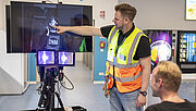 Sicherheitsmanager André Wilhelmi deutet auf den Bildschirm der UV-Kamera. Dieser zeigt einen Mitarbeiter des Amazon-Sortierzentrums, der einen Teil seines Gesichts mit Sonnencreme eingecremt hat. Die eingecremte Gesichtshälfte wird schwarz dargestellt. Der Mitarbeiter sitzt ein paar Meter vor dem Bildschirm und hört den Erklärungen von André Wilhelmi zu. 