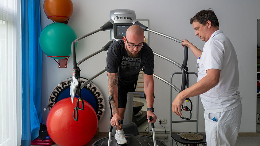 Moritz Sontheimer in der Physiotherapie der BGU Murnau. Er lehnt sich nach vorne und stützt sich auf seine Gehhilfen, rechts an seiner Seite steht Physiotherapeut Tobias Bergmüller. 