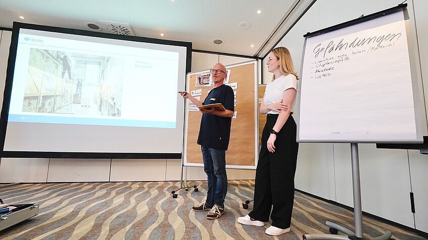 Ein Mann und eine Frau stehen vorne im Seminarraum. Im Hintergrund ist eine große Leinwand mit einer Präsentation zu sehen, daneben ein Flipchart mit der Überschrift „Gefährdungen“. Auf dem Flipchart sind Stichpunkte notiert, unter anderem „Umstürzen von Ware/Material“, „Staplerunfälle“ und „Lasten“. Die Teilnehmenden sitzen im Raum.